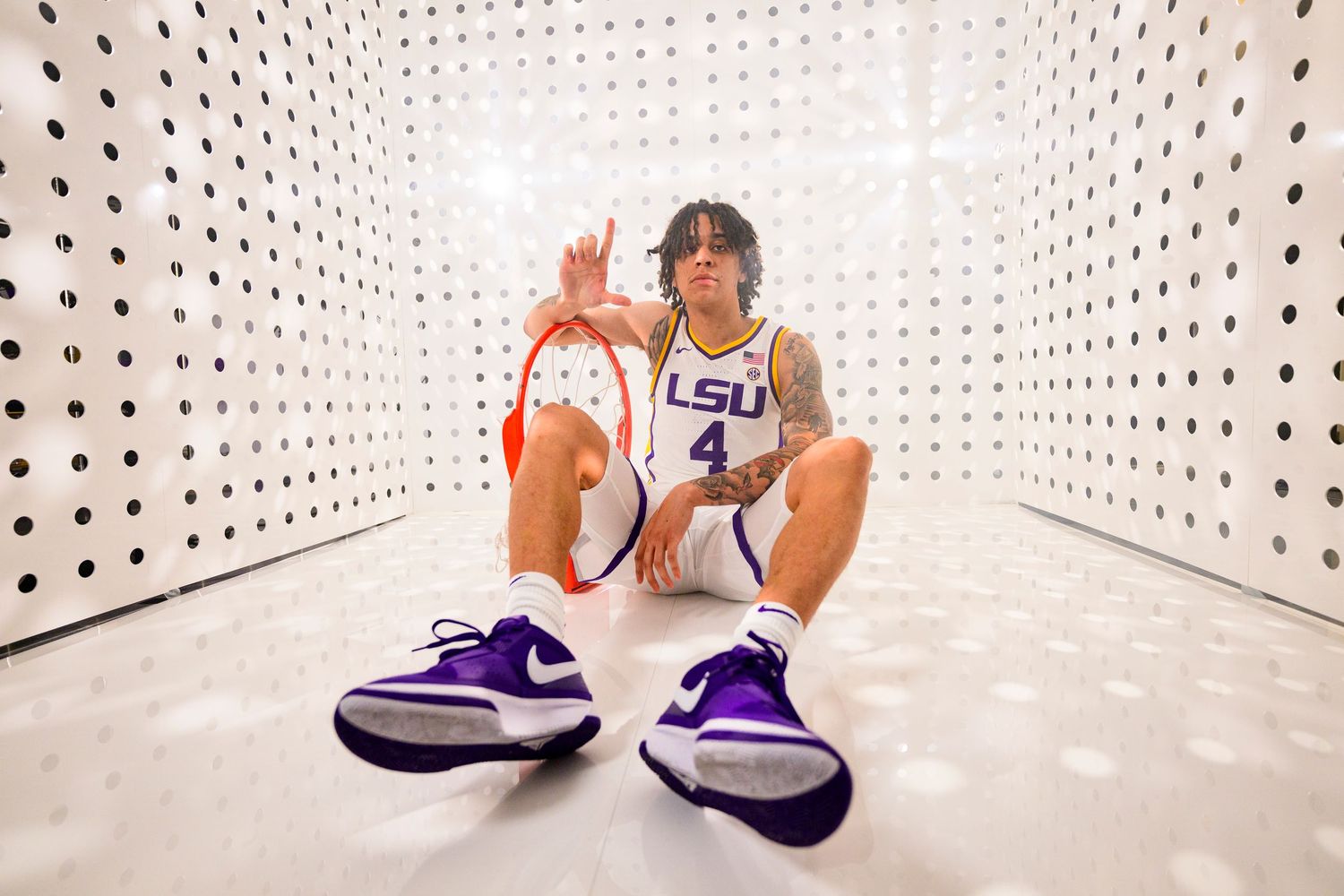 LSU athlete sits in white pegboard hallway wearing uniform with bright purple Nike sneakers in front.