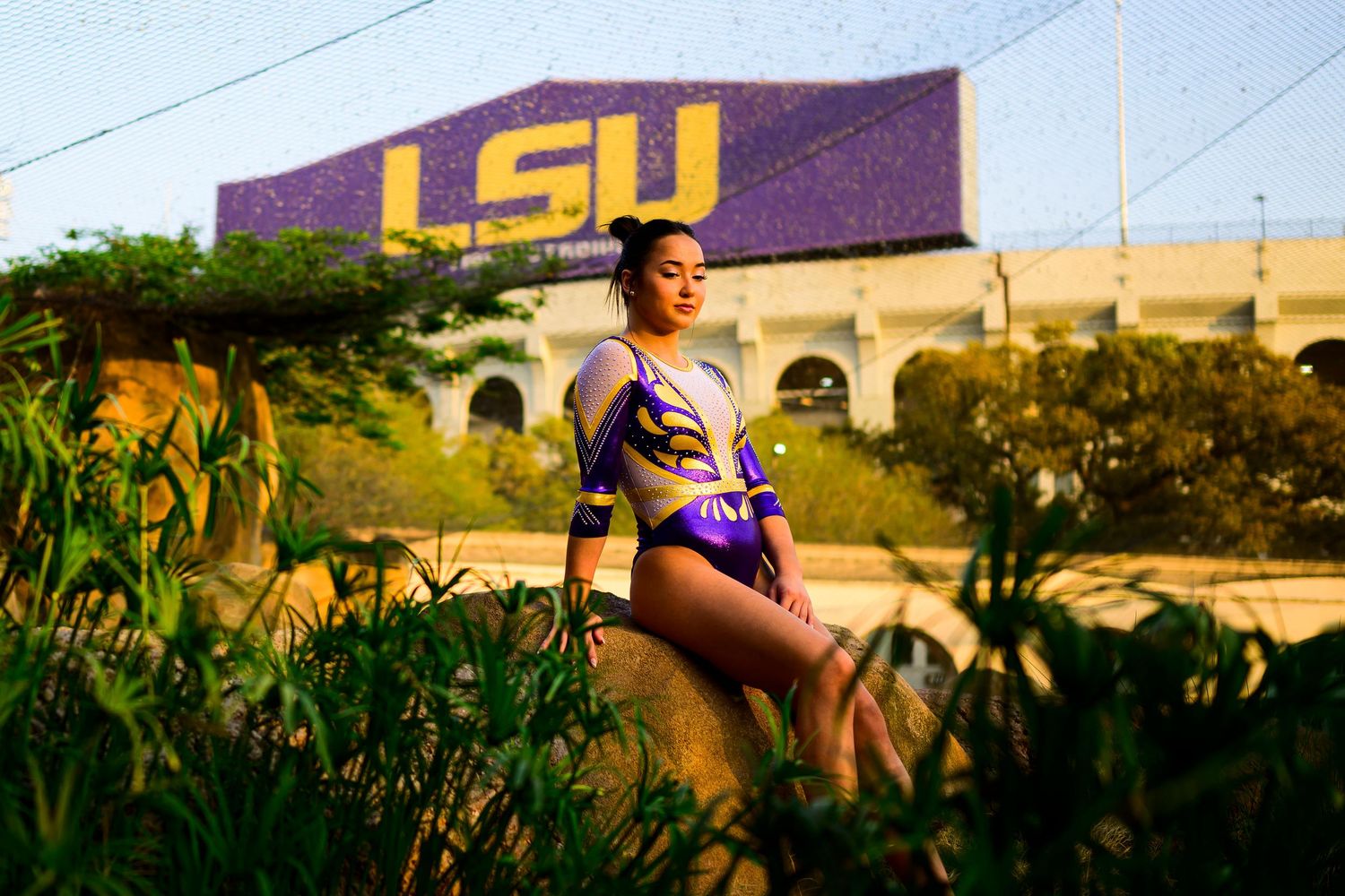 Athlete in purple LSU gymnastics leotard poses in grass field with stadium and LSU banner in background.