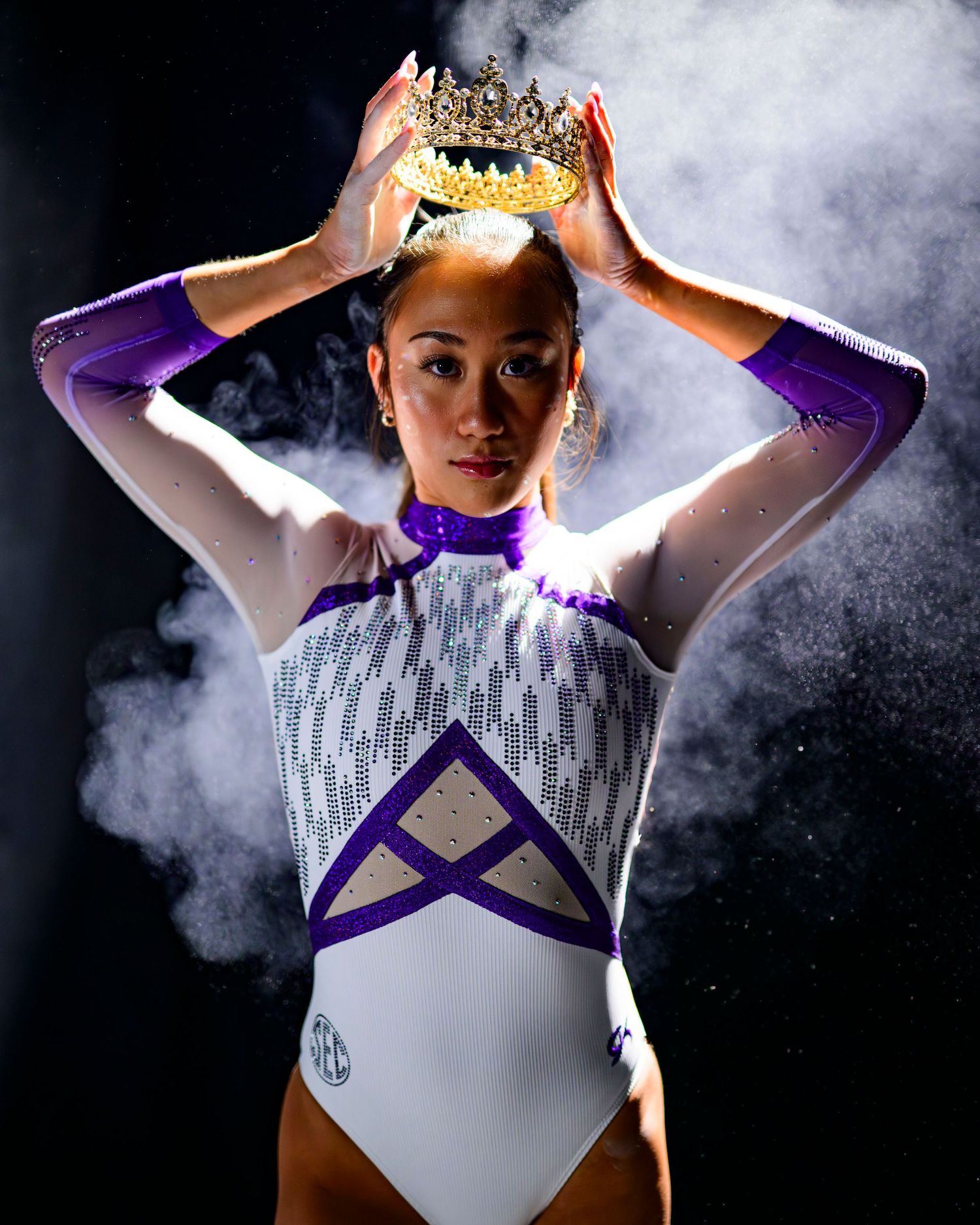 Artistic portrait of a gymnast in white and purple leotard posing with a crown amid dramatic smoke effects.