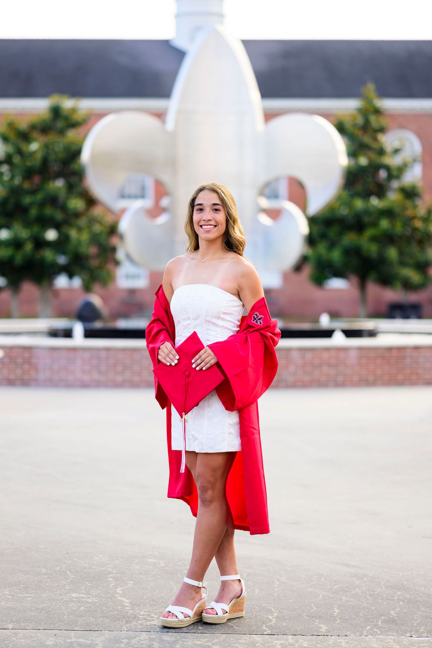 Graduate poses in white dress and red stole in front of fleur-de-lis campus monument.