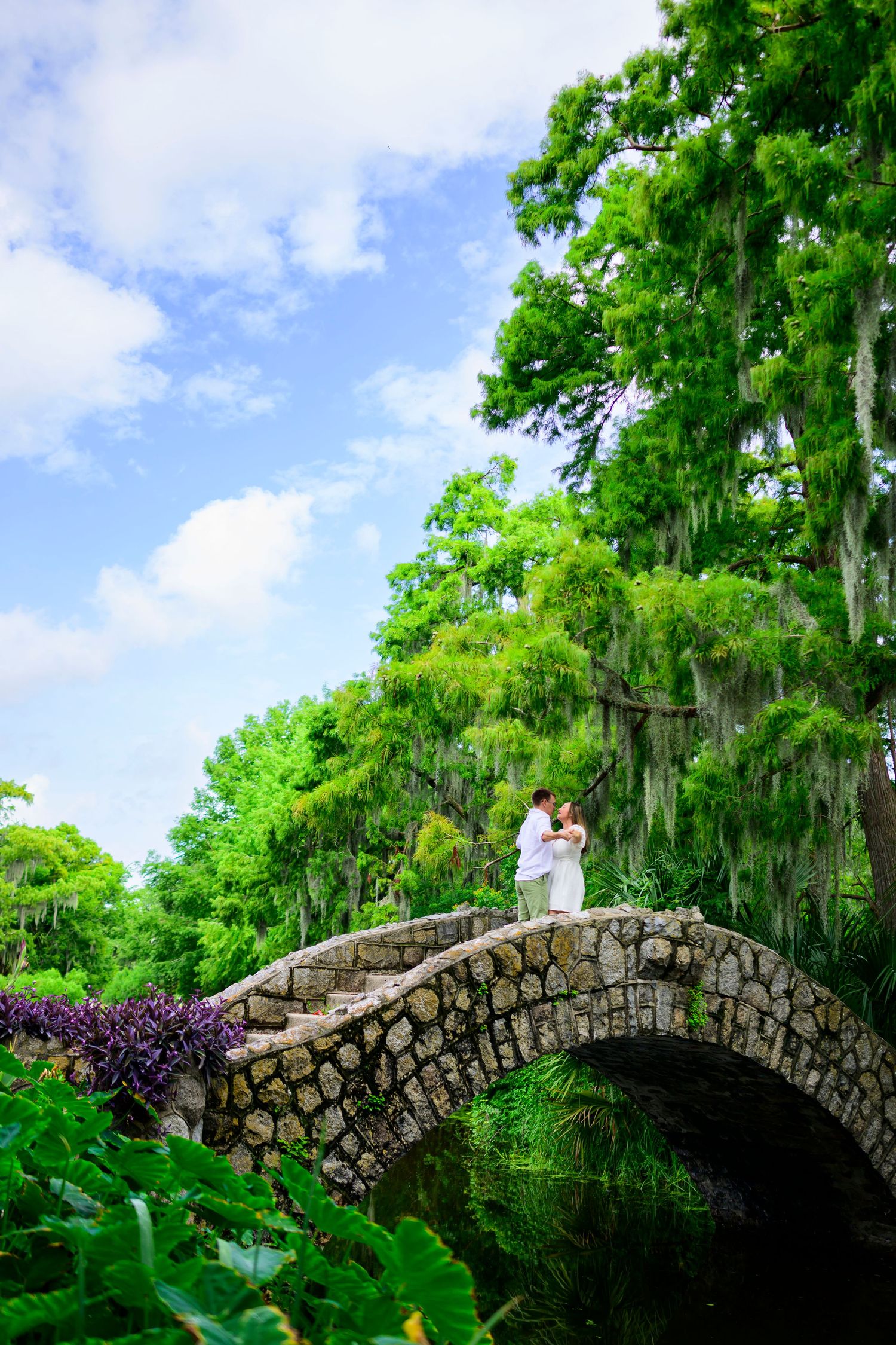 Couple standing on a stone arch bridge surrounded by lush gardens and Spanish moss.