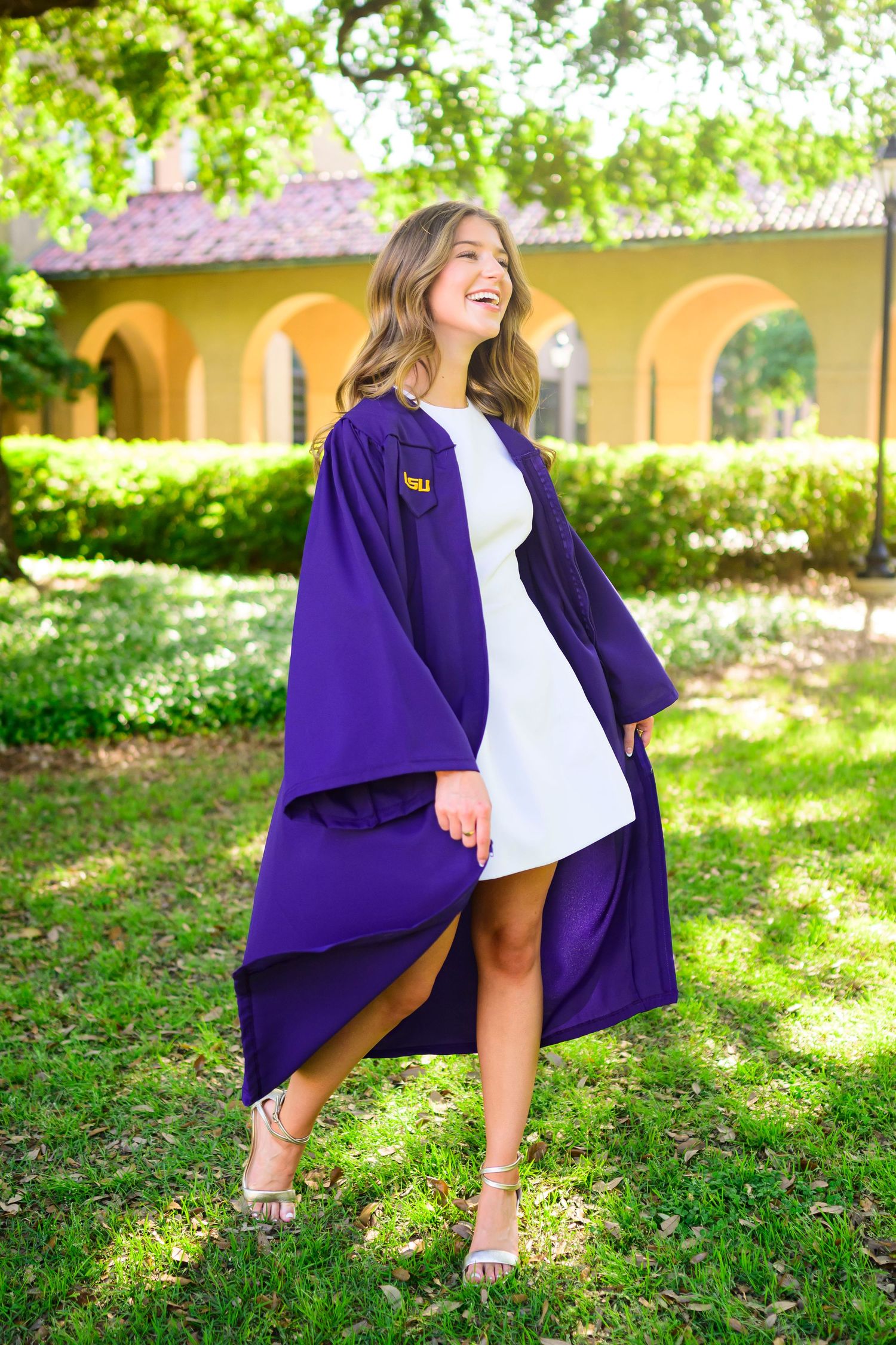 Graduate in purple LSU graduation gown poses in front of Spanish-style campus architecture.