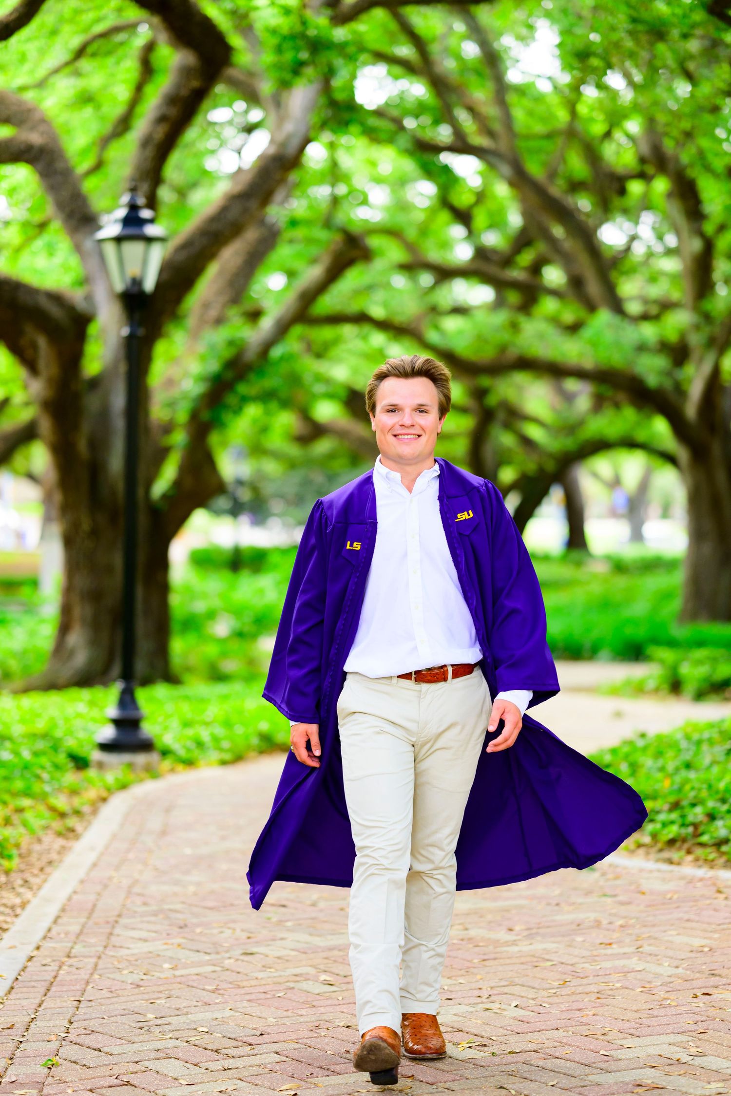 Graduate in purple graduation gown walks along tree-lined brick path.