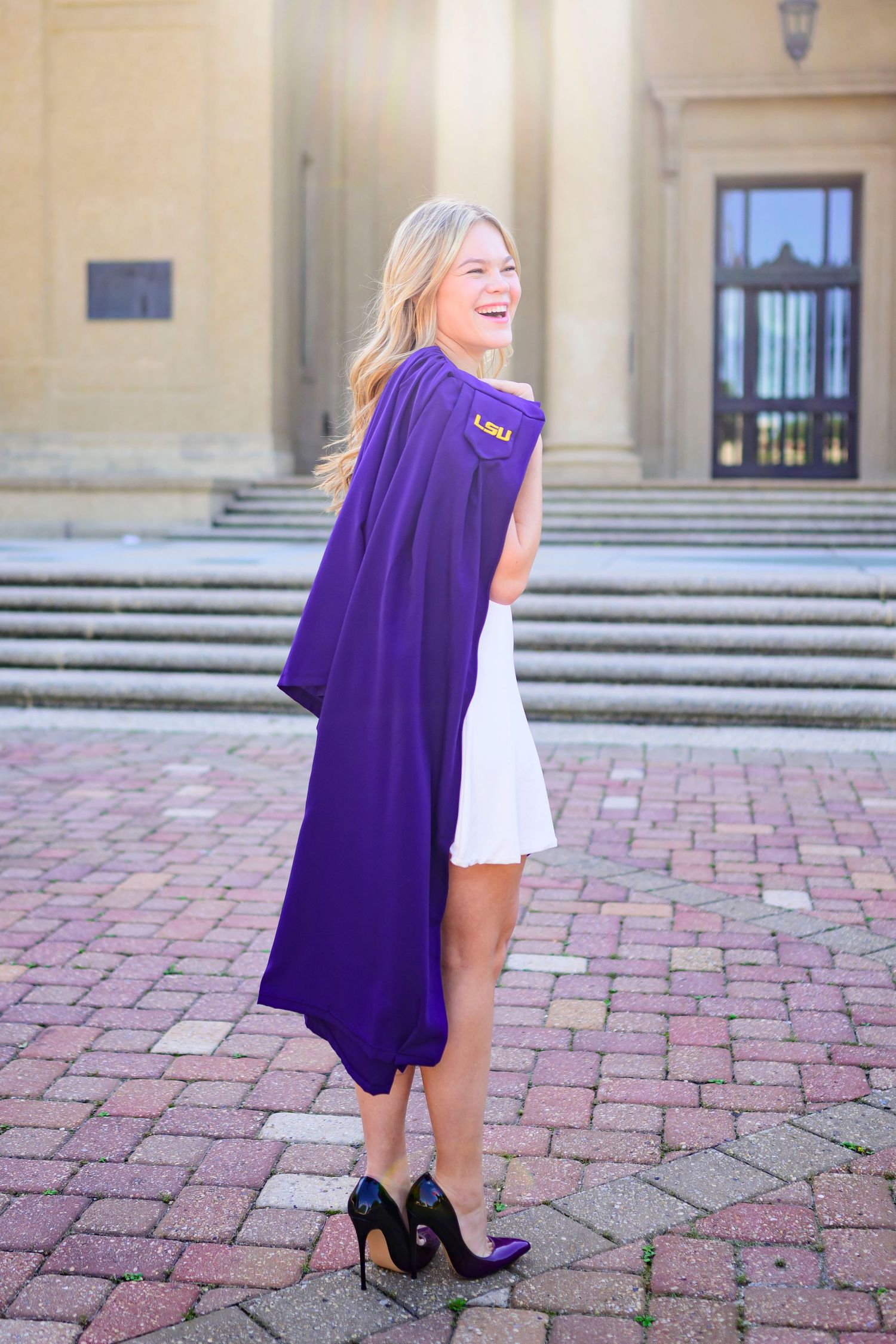 Graduate in purple academic regalia poses on brick courtyard with classic architecture.