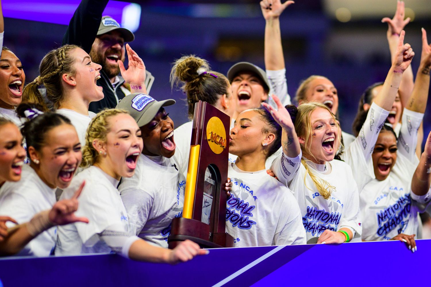 Team celebrates with championship trophy as confetti falls during college sports victory celebration.