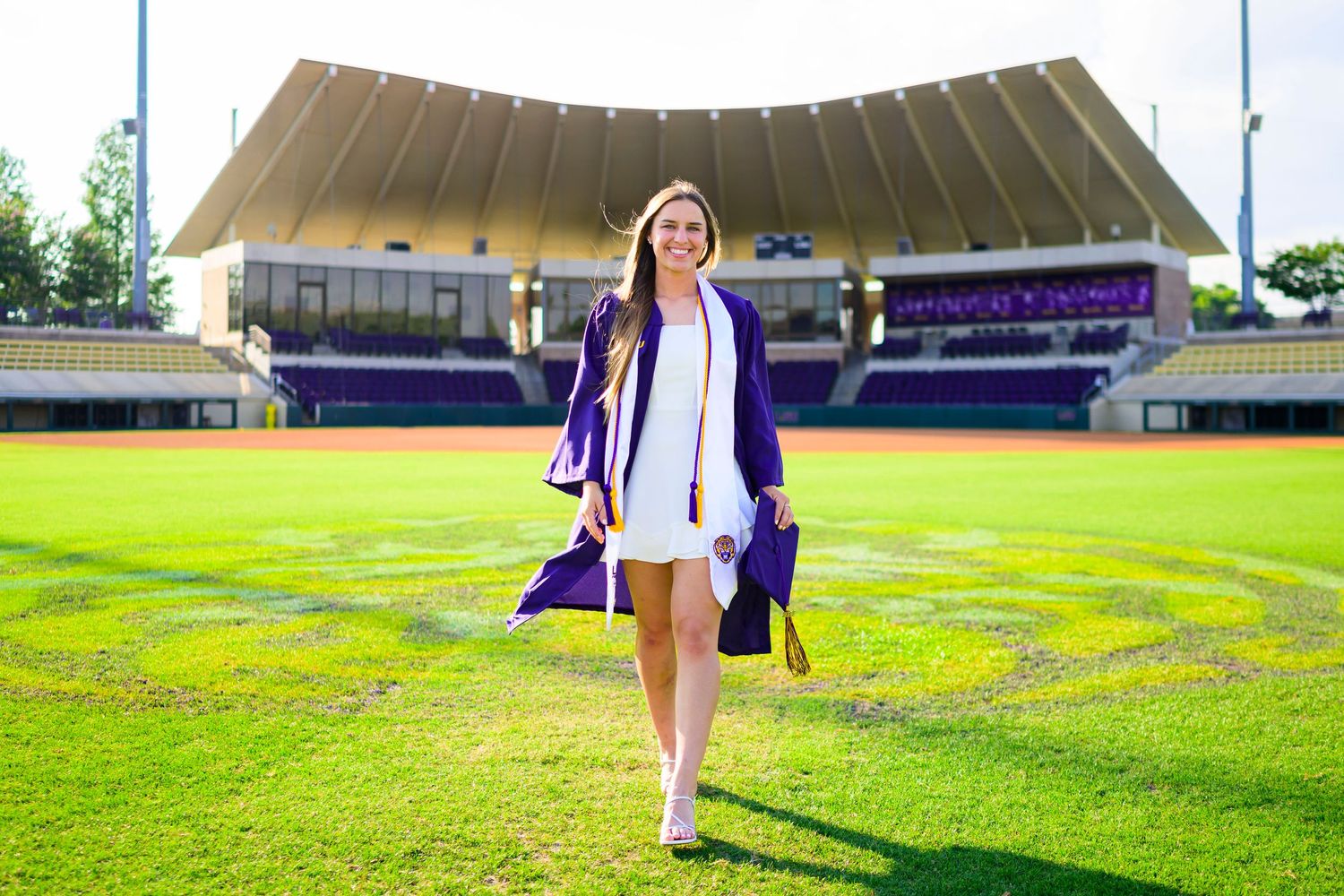 Graduate walking across sports field in purple graduation gown at outdoor stadium.