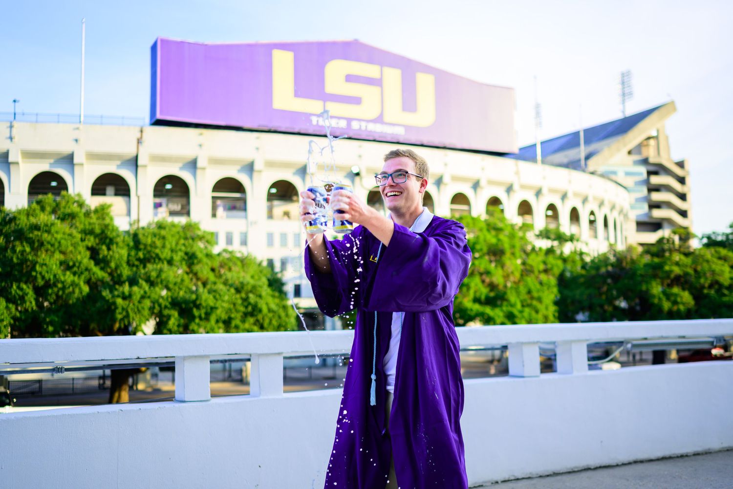 Graduate in purple gown celebrating in front of LSU stadium with arms raised.