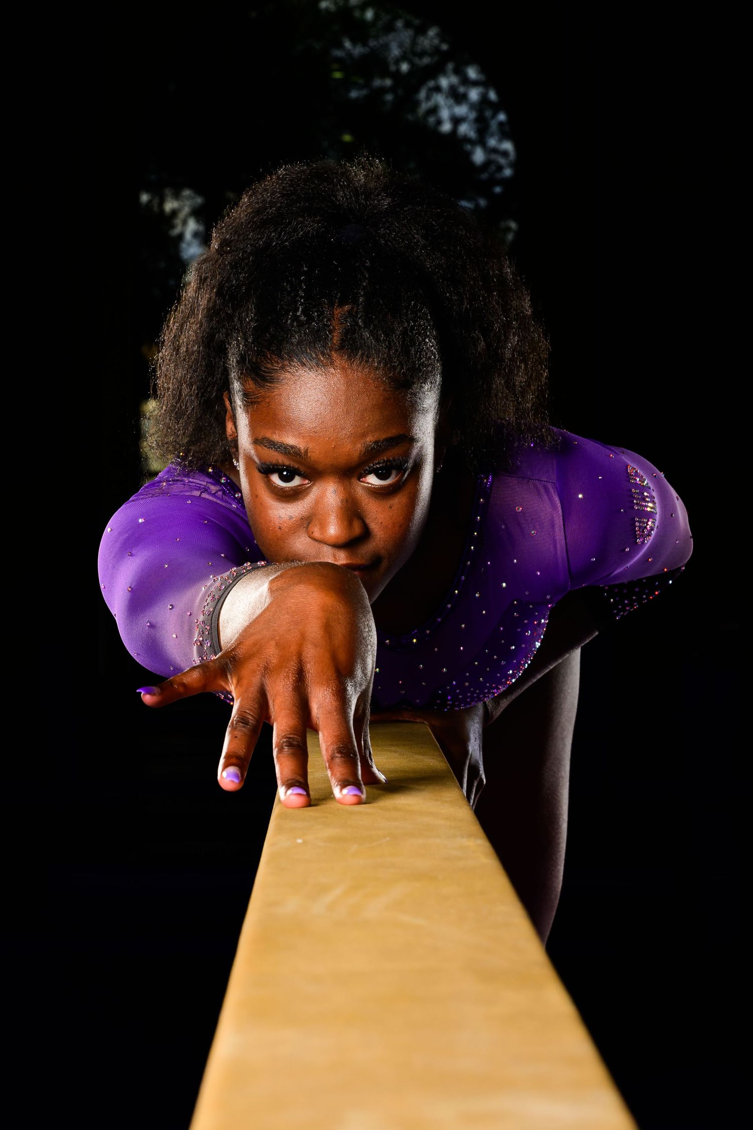 Gymnast in sparkling purple leotard poses on balance beam against black background.