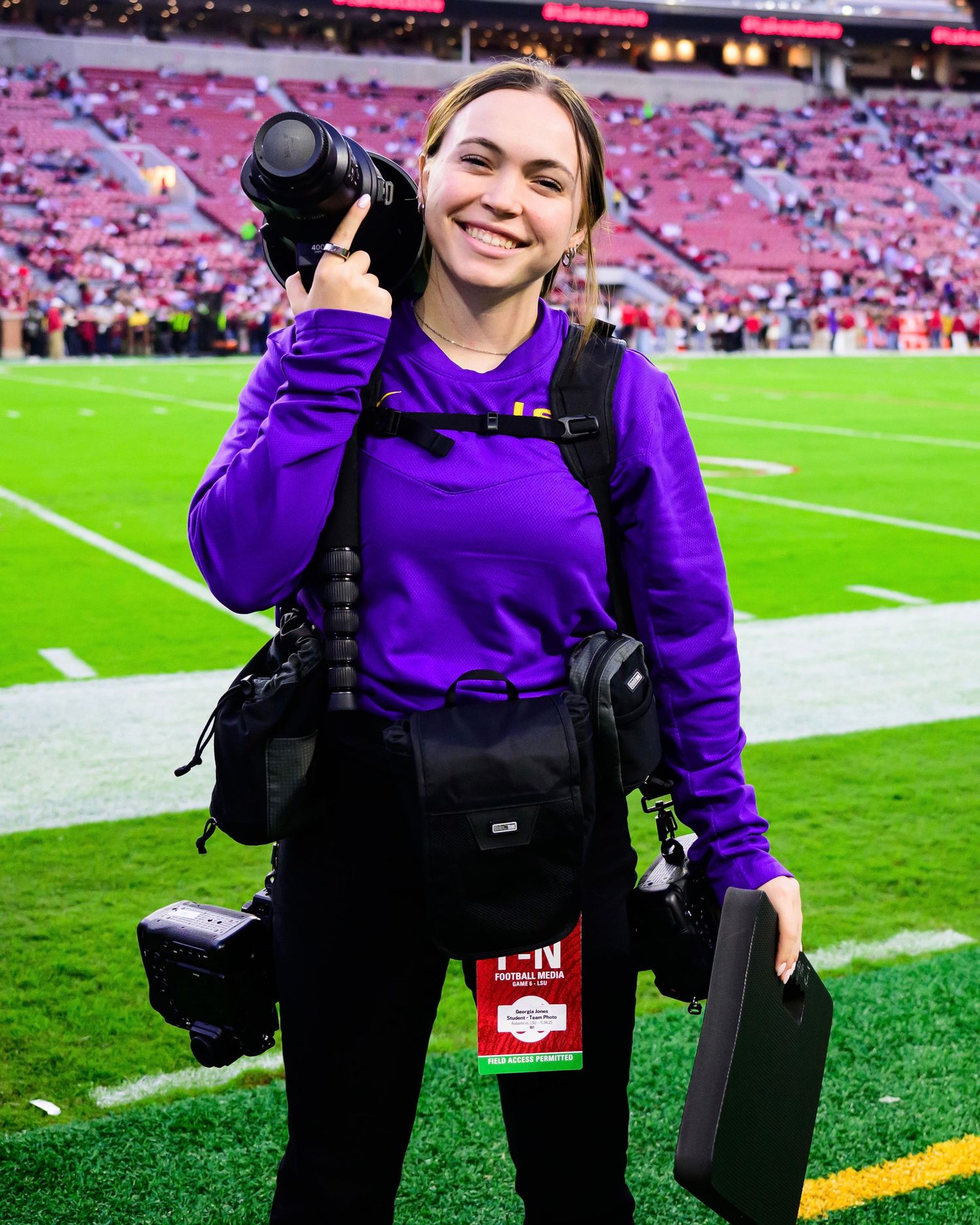 Sports photographer in purple sweatshirt with professional camera gear on football field.