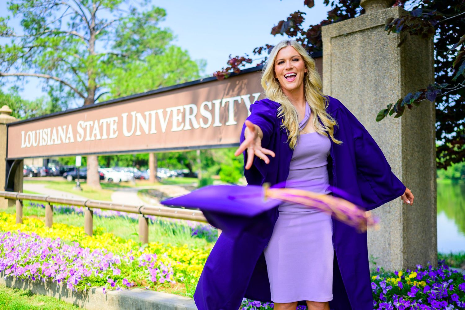 Graduate in purple regalia poses by Louisiana State University entrance sign.