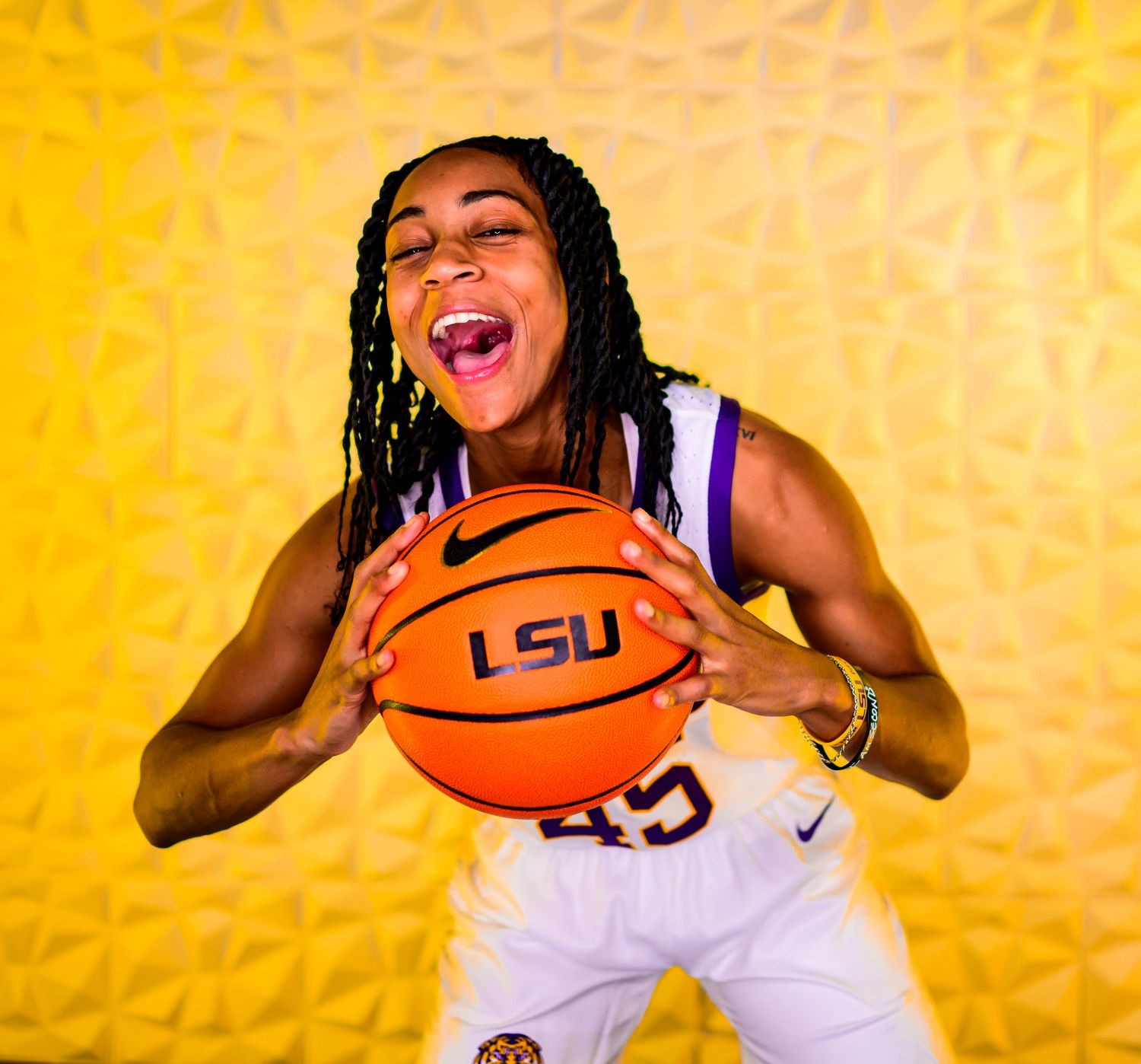 Basketball player in LSU uniform holds Nike basketball while laughing against vibrant yellow backdrop.