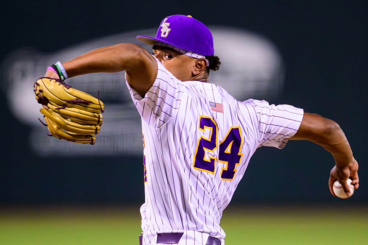 A baseball pitcher in a white and purple pinstriped uniform with number 24 delivers a pitch during a nighttime game.