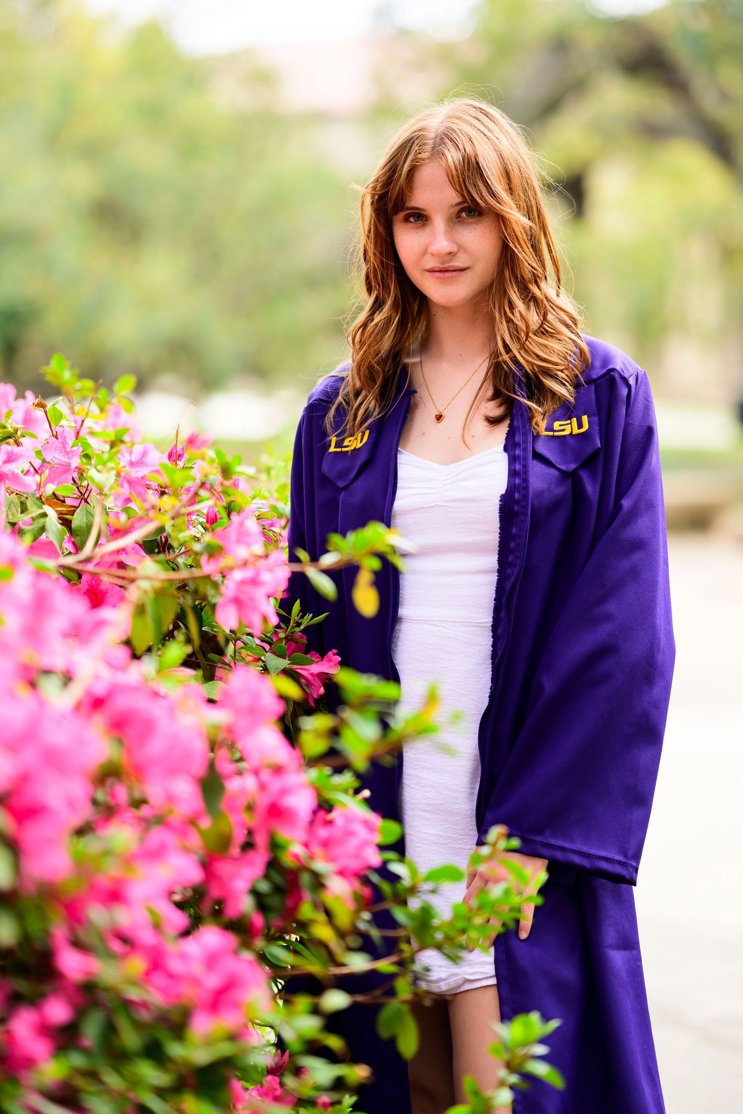 Student in purple LSU academic regalia poses by blooming pink flowers.