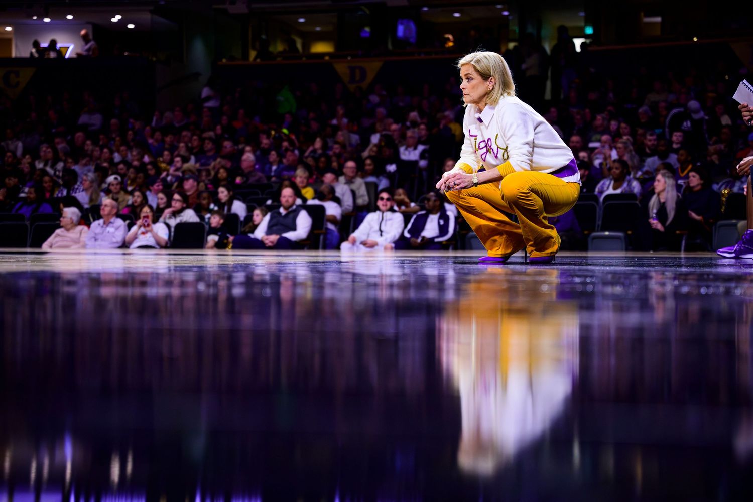 Performer in yellow outfit crouches on illuminated stage with reflection in dark venue.