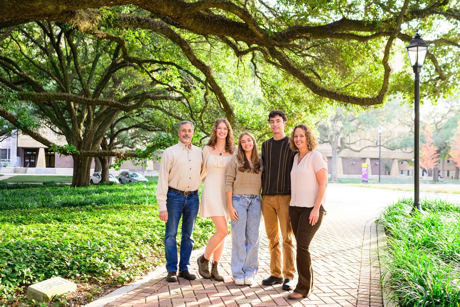 Group standing on a brick pathway under sprawling oak trees in a lush garden setting.