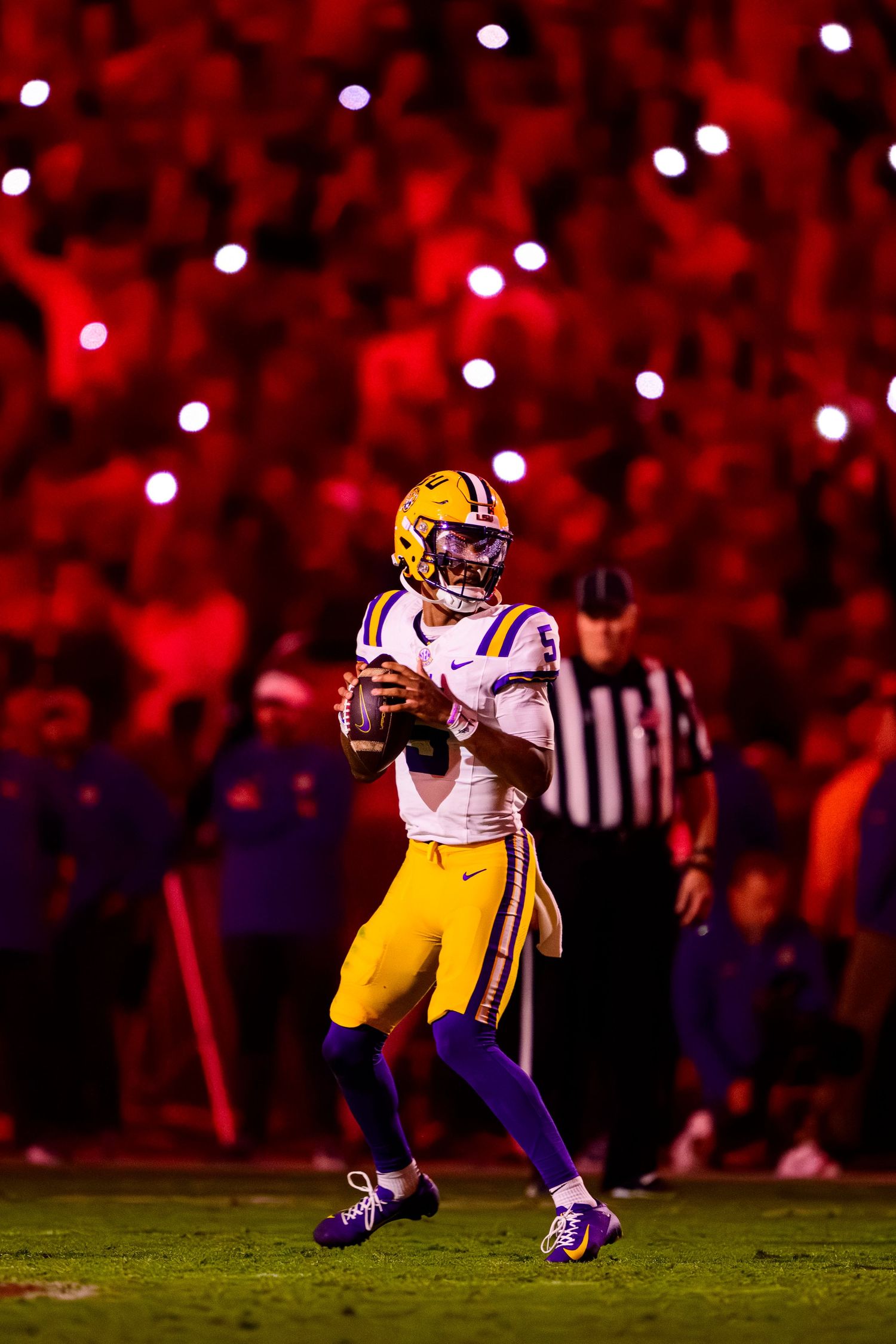 Football player in yellow LSU uniform drops back to pass under dramatic red stadium lighting.