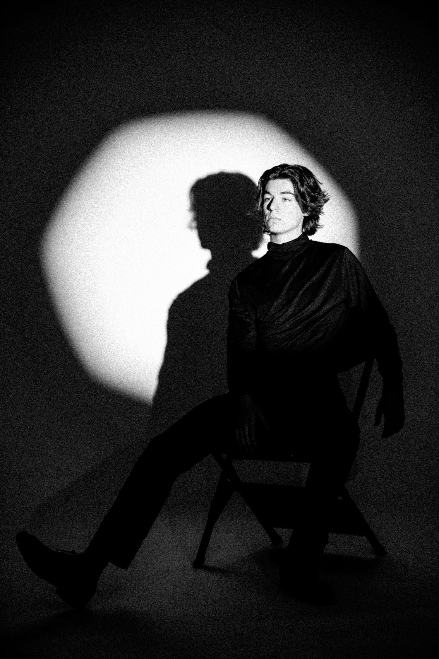 Artistic black and white portrait of figure seated in dramatic lighting with shadow cast on circular background.
