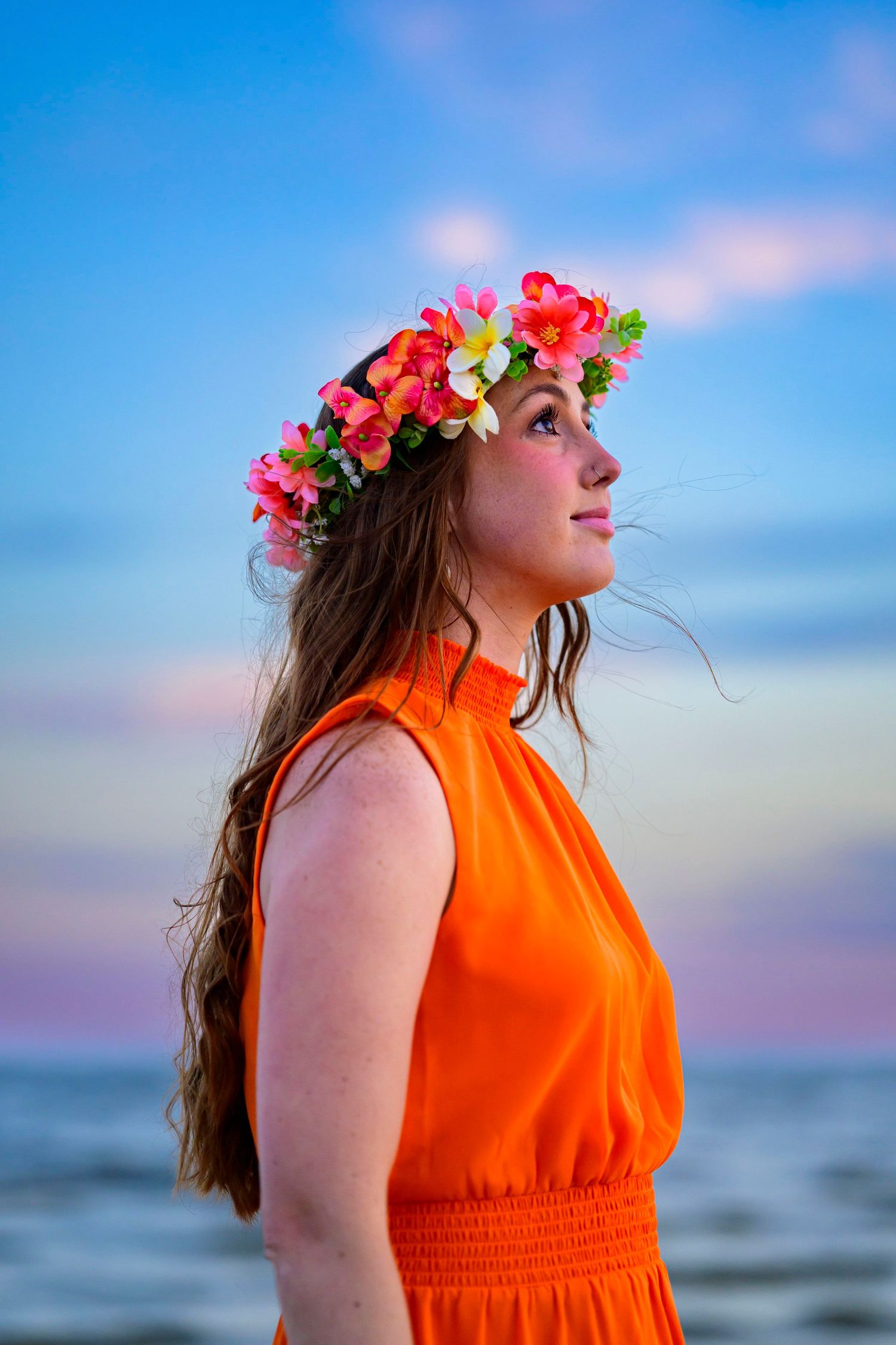 Woman in orange dress wearing floral crown poses against sunset sky at beach.