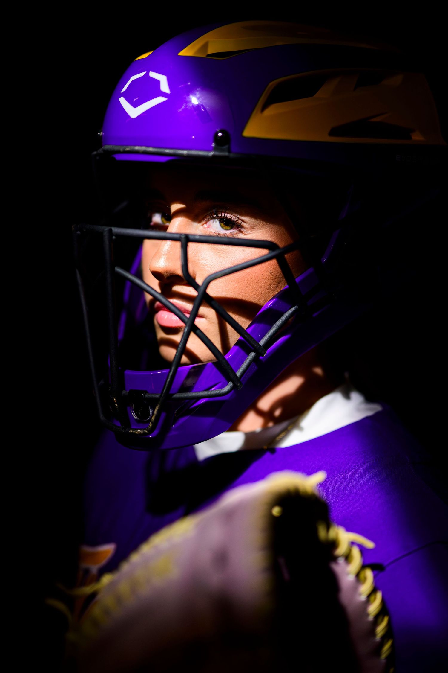 Close-up shot of a baseball catcher wearing a purple and gold helmet with protective face mask in dramatic lighting.