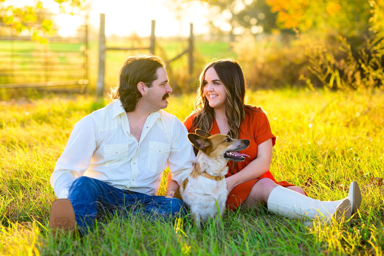 Couple sitting in golden sunlit field with their white dog during sunset.