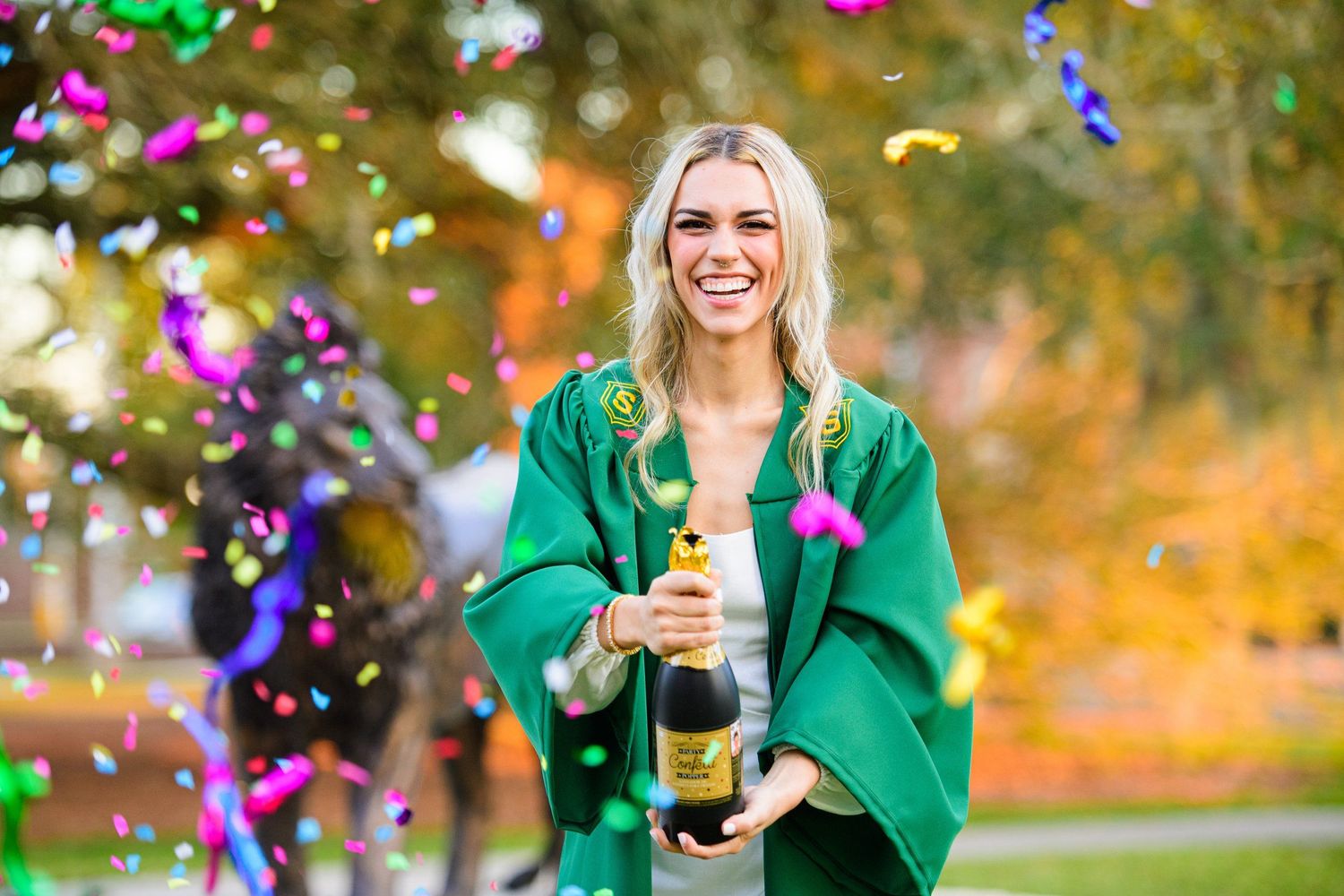 Graduate in emerald green gown celebrates with confetti and champagne outdoors.