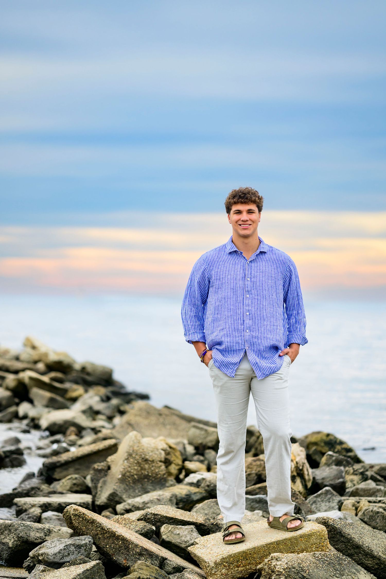 Casual coastal portrait with blue button down shirt and khakis on rocky beach at sunset.