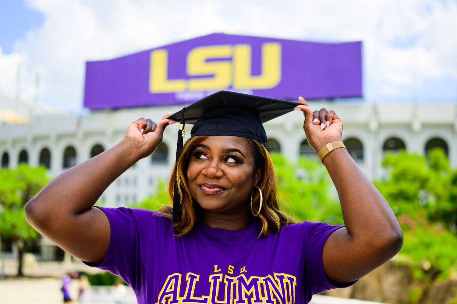 Graduate adjusting cap in front of LSU stadium while wearing purple alumni shirt.