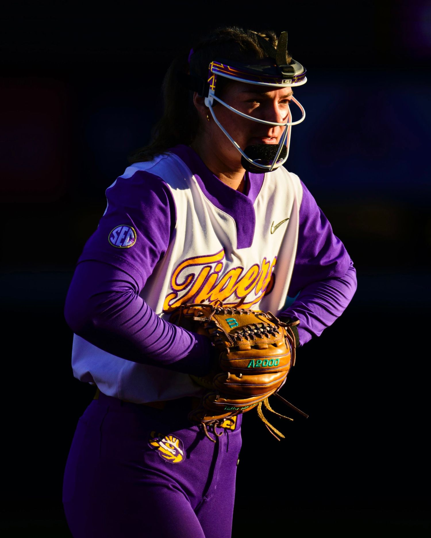 Softball player in purple LSU uniform and protective mask prepares to pitch.
