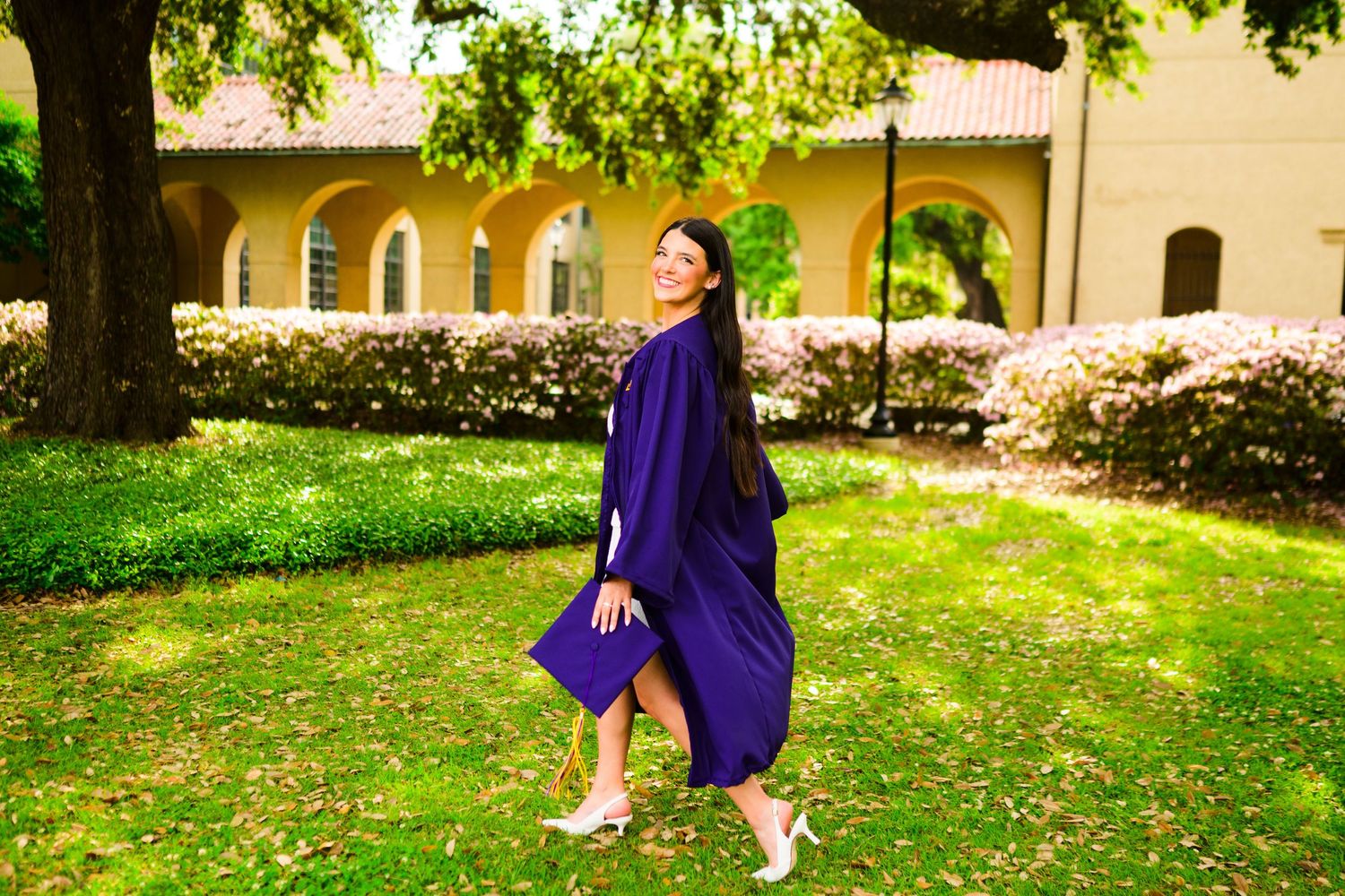 Woman in purple graduation gown smiling outdoors on green campus lawn with trees and building backdrop.