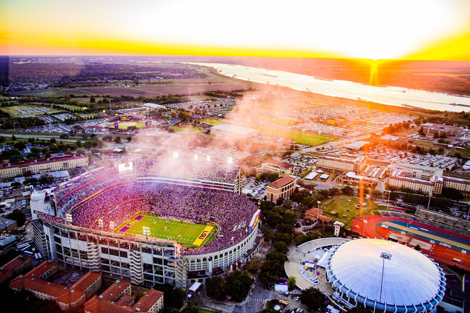 Aerial view of sports stadium lit up at sunset with colorful sky and dome in background.