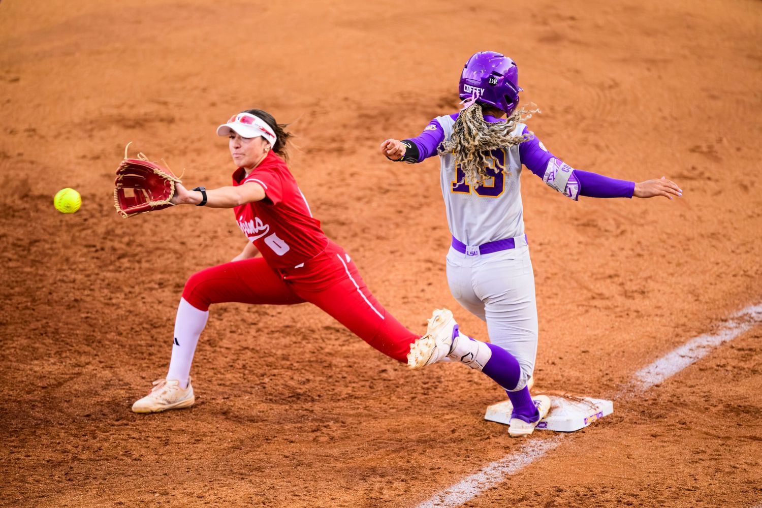 Softball players compete at first base during intense game action on dirt infield with one sliding into base.
