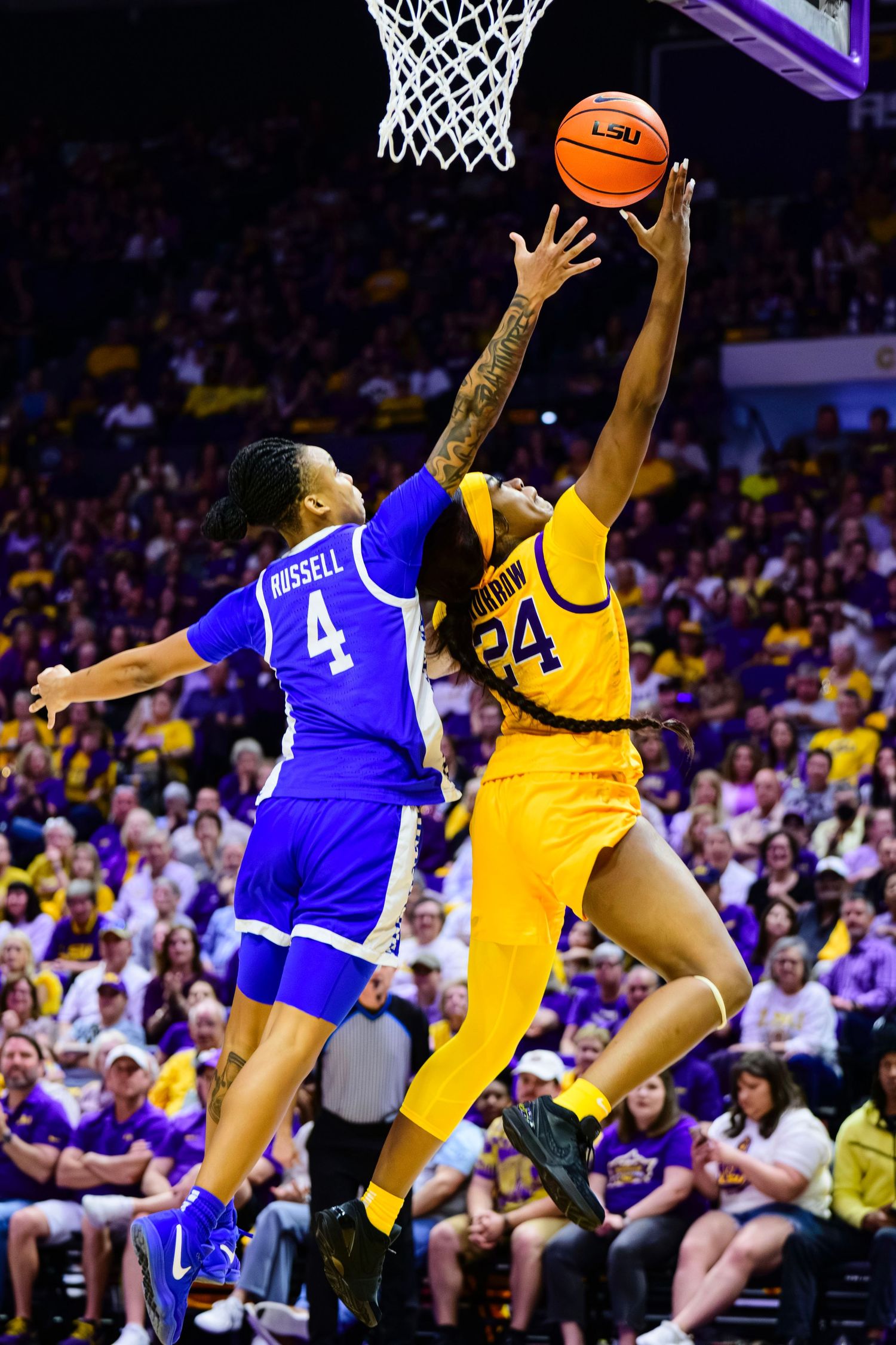 Two basketball players leap high for the ball during an intense college game between teams in blue and yellow uniforms.
