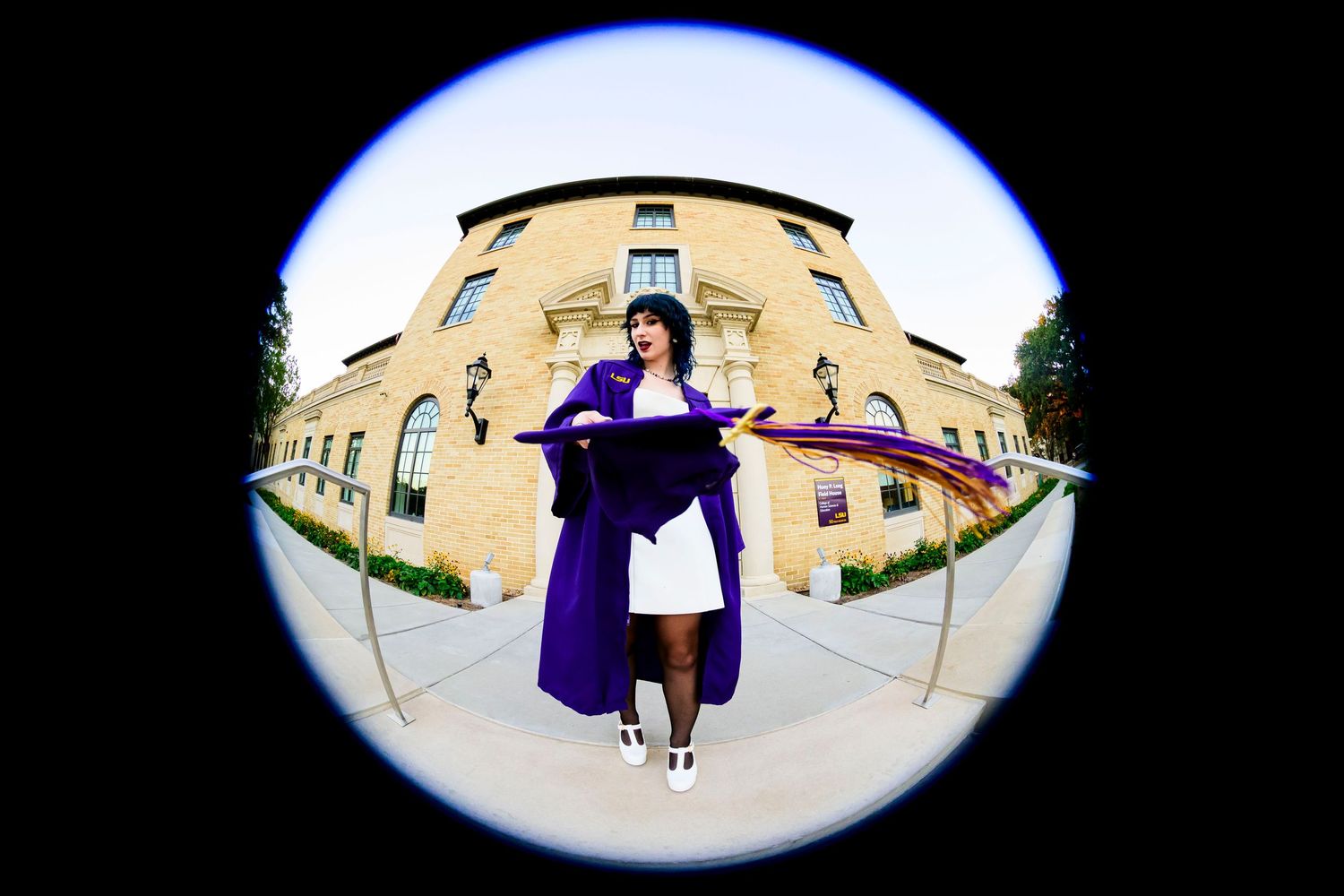 Fisheye lens view of graduate in purple gown standing in front of building.
