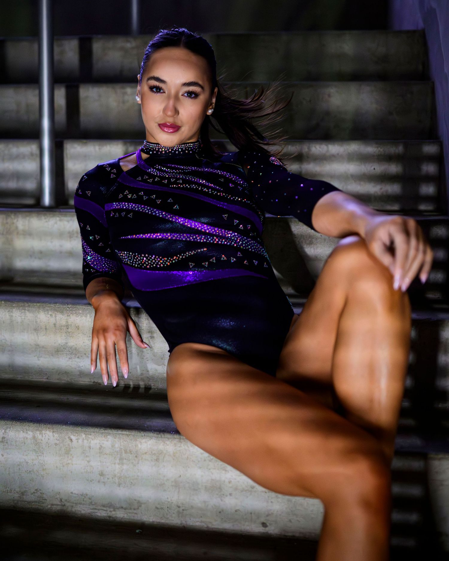 Dancer in sparkly purple and black leotard poses dramatically on concrete steps in moody lighting.