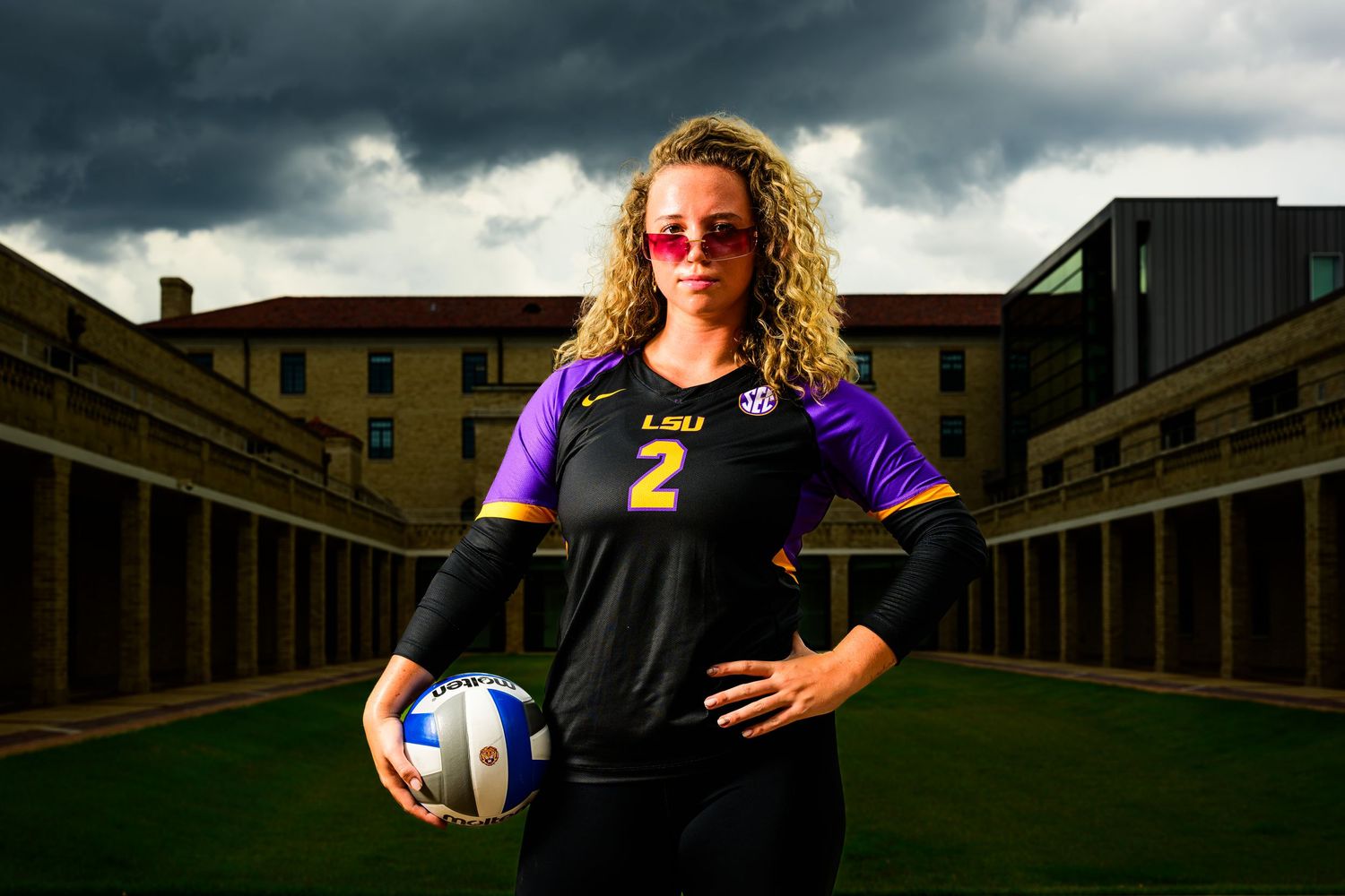 LSU volleyball player wearing jersey number 2 poses with volleyball under dramatic stormy sky on campus courtyard.