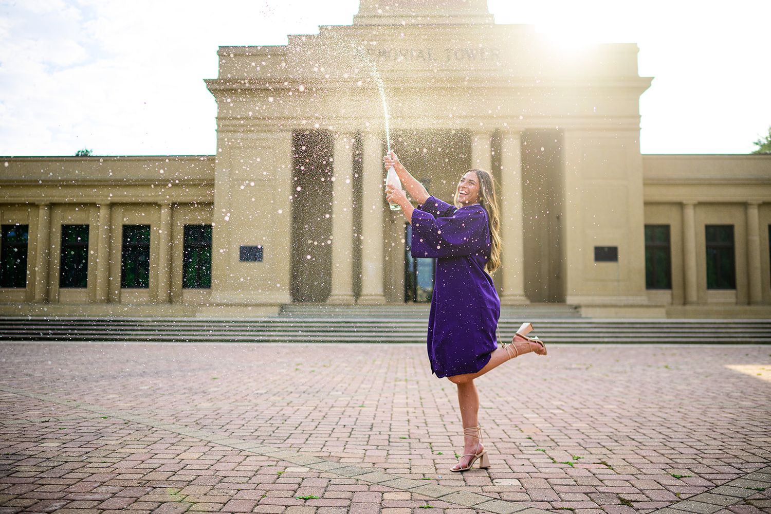 Graduate celebrates in a purple dress twirling on brick pavement in front of historic building architecture.