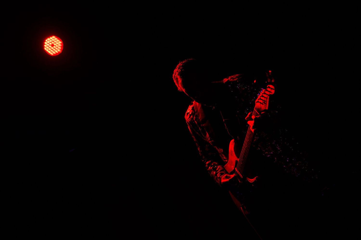 A guitarist performs on stage bathed in dramatic red lighting against a dark background.