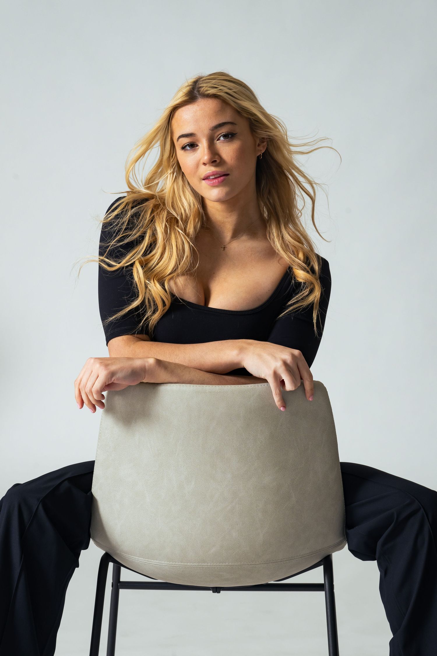 Fashion portrait of woman in black outfit posing with modern chair against white background.