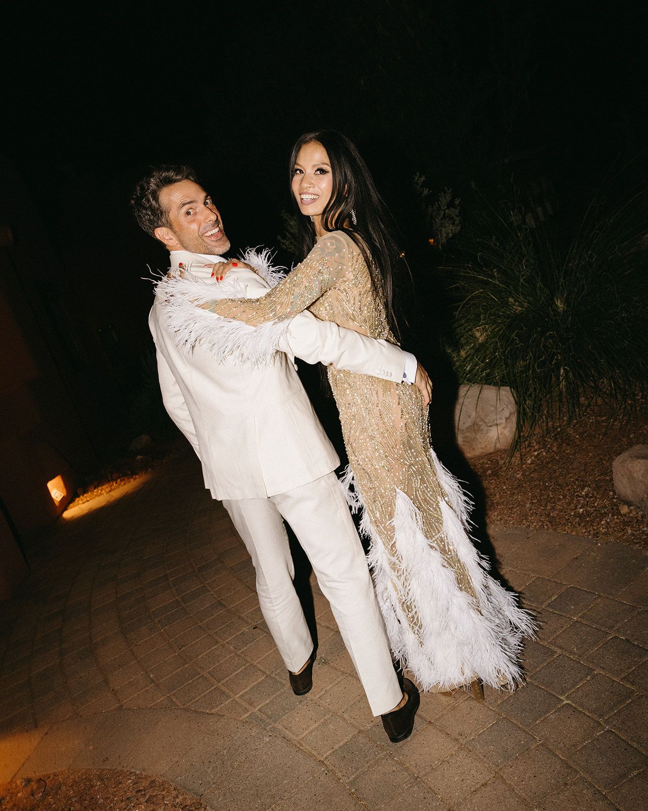 Two people in white formal attire dancing joyfully on a paved outdoor patio at night.