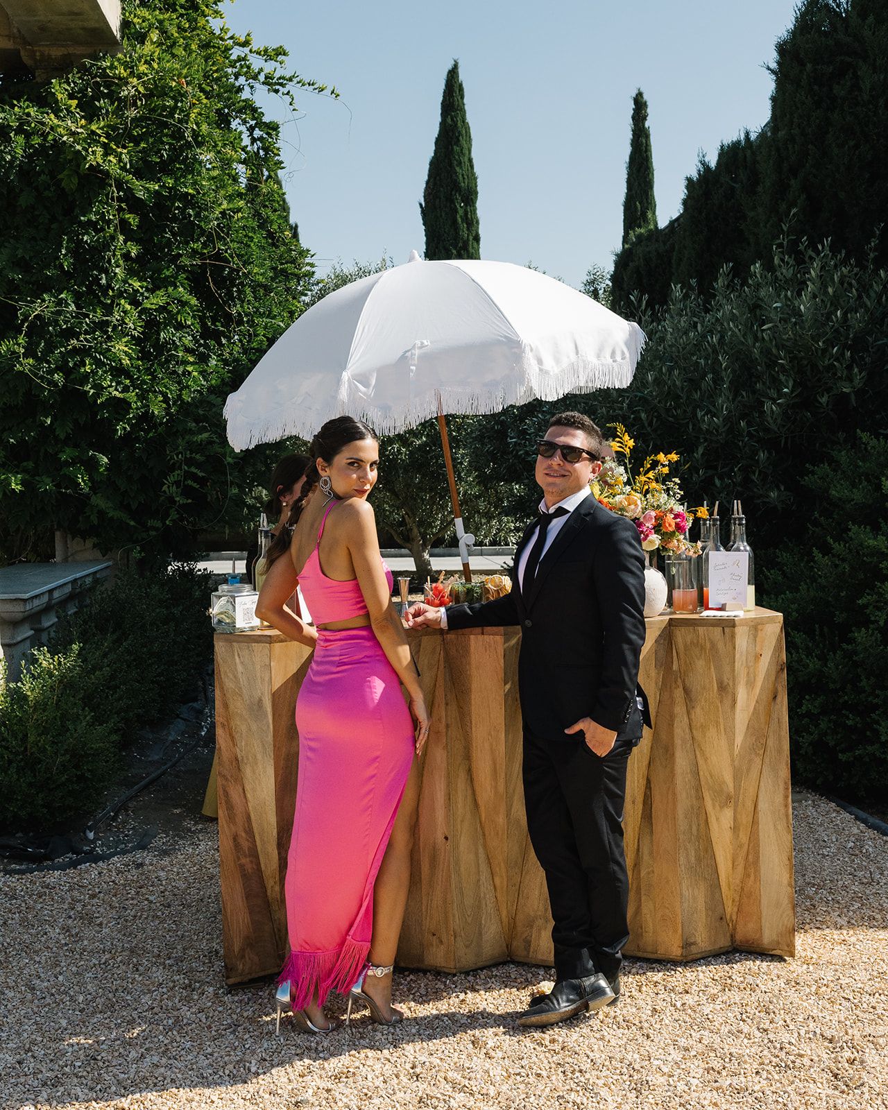 Wedding bar setup with white umbrella and wooden crates in garden setting with cypress trees.