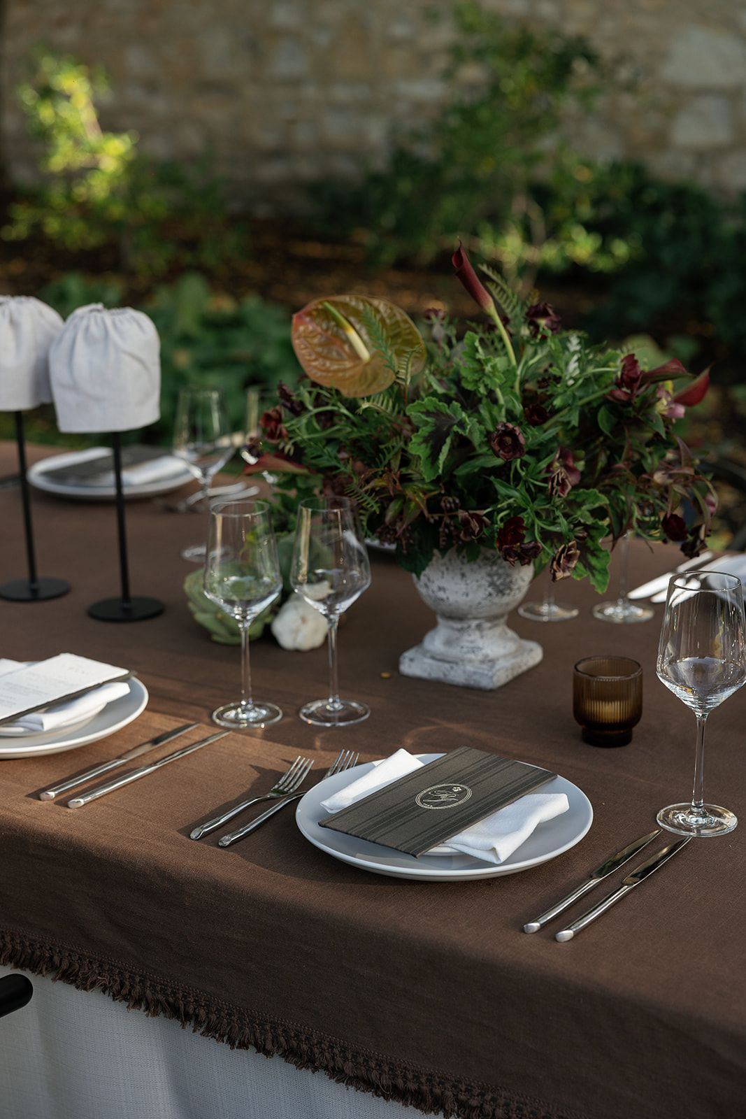 Elegant table setting with brown tablecloth, wine glasses, and fresh greenery centerpieces at upscale dining event.