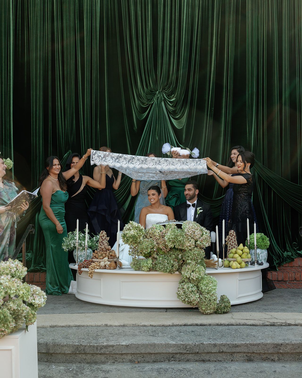 Wedding party members strike fun poses while lounging on a curved white bench with green floral decorations at an indoor venue.
