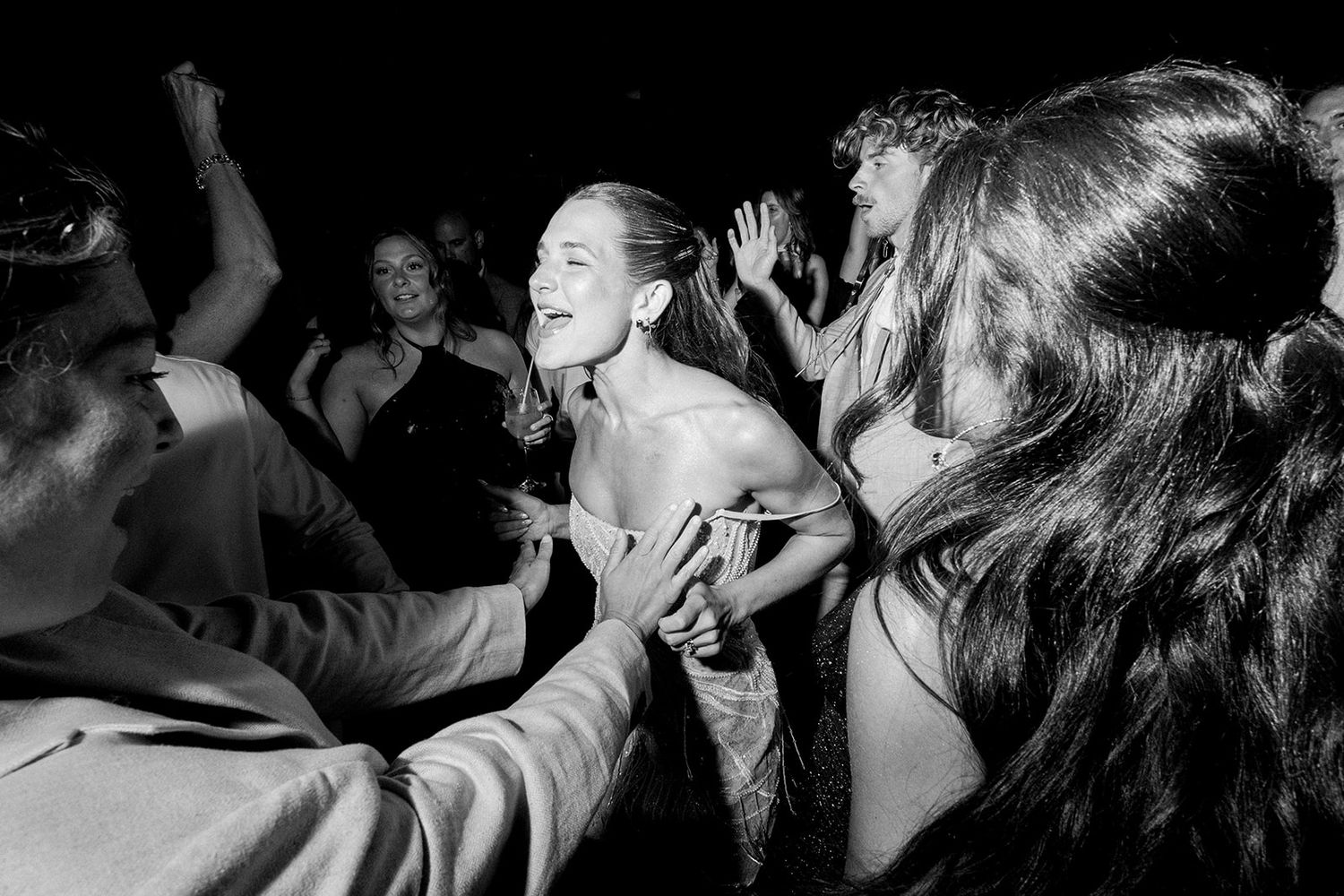 Black and white photo of a smiling woman dancing joyfully at a lively party surrounded by guests.