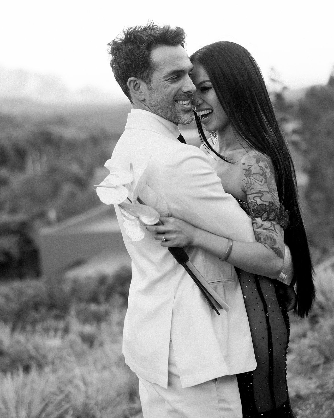 Black and white candid romantic photo of a couple embracing outdoors against a scenic landscape backdrop.