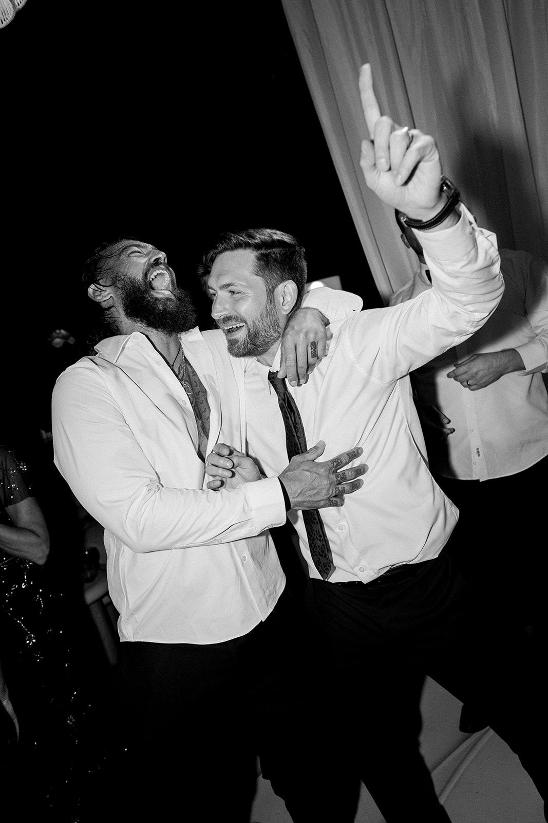 Two bearded men in white dress shirts laugh and dance joyfully at a formal evening event.