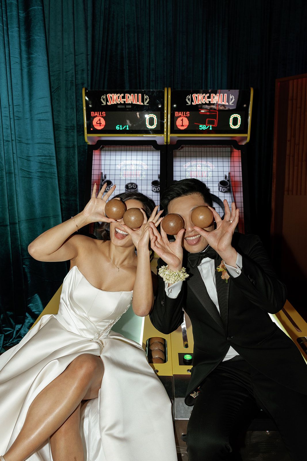 Bride and groom playfully hold skee-ball game balls over their eyes at a fun wedding reception.