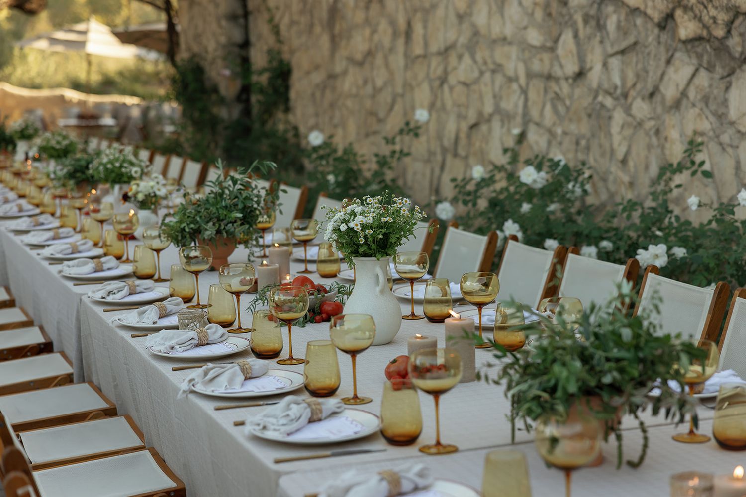 Elegant outdoor dining table set with amber glassware, white plates and rustic greenery centerpieces along stone wall.