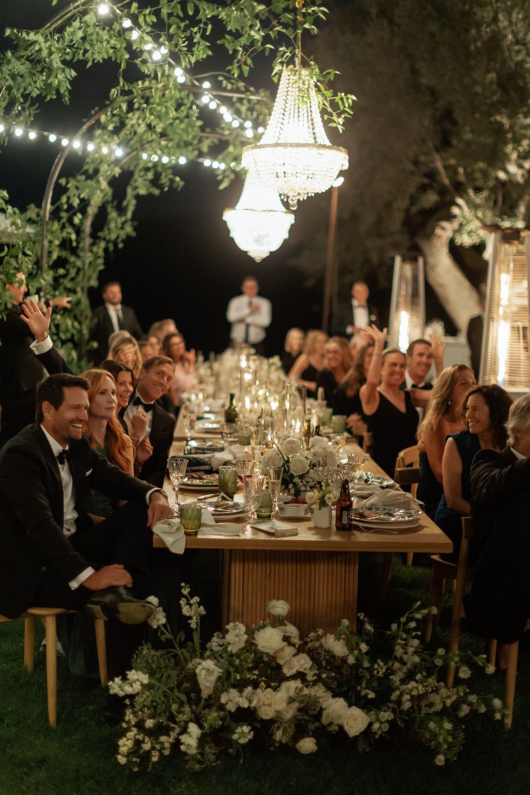 Elegant outdoor wedding reception at night with crystal chandeliers and guests seated at long tables beneath trees.