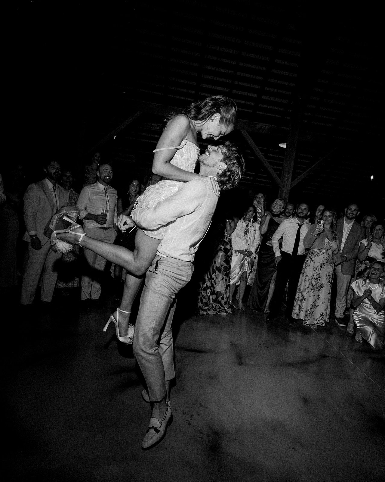 Black and white photo of groom lifting bride during first dance at wedding reception with guests watching.
