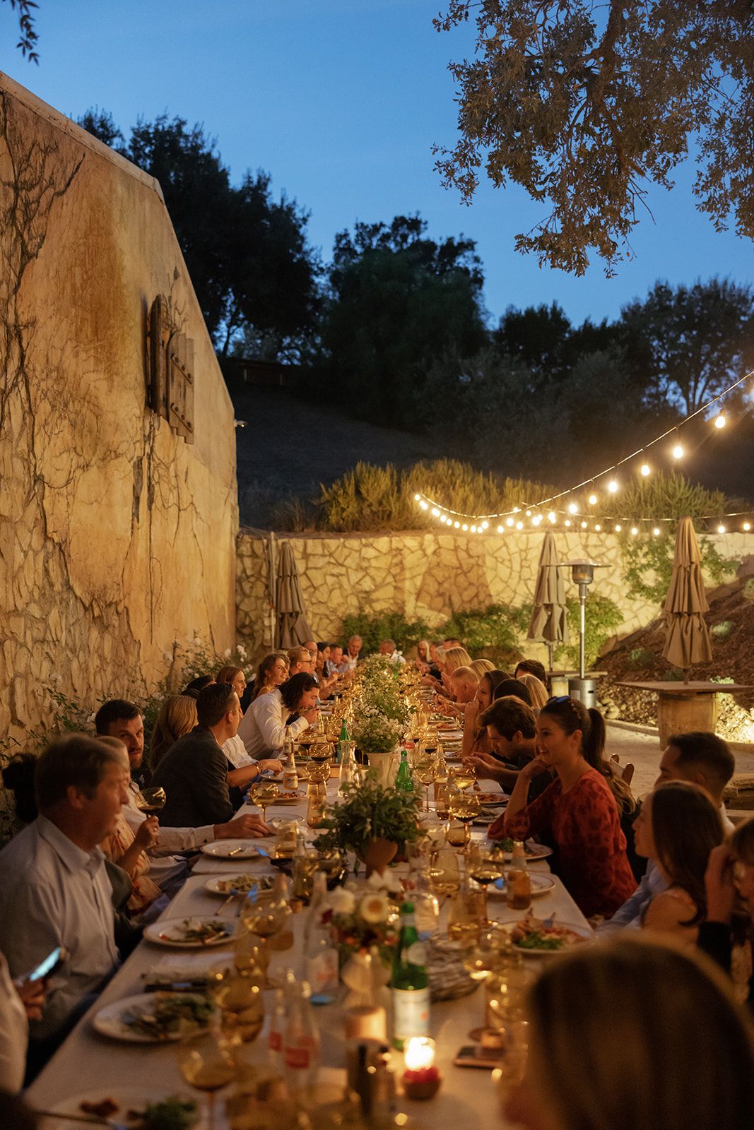 Guests gather for an elegant outdoor dinner party under string lights at dusk with long tables and floral centerpieces.